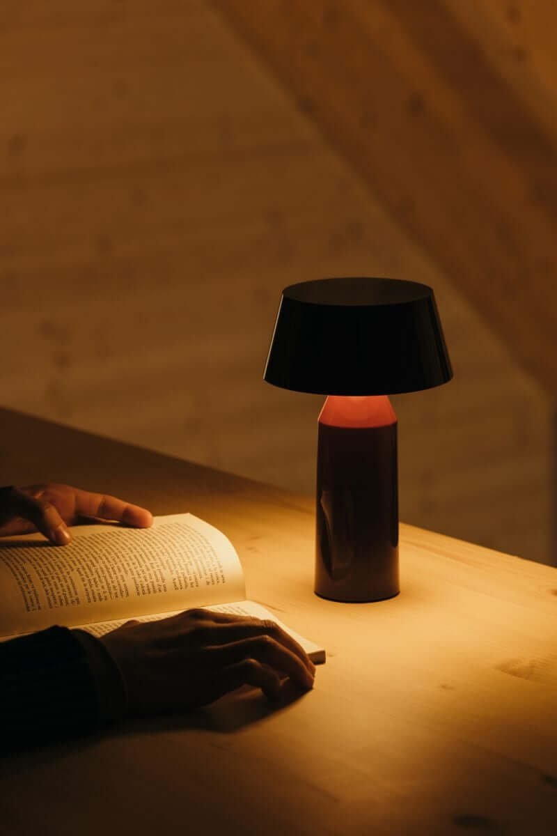 Marset Bicoca table lamp in black and red that illuminates a book on a wooden table. 